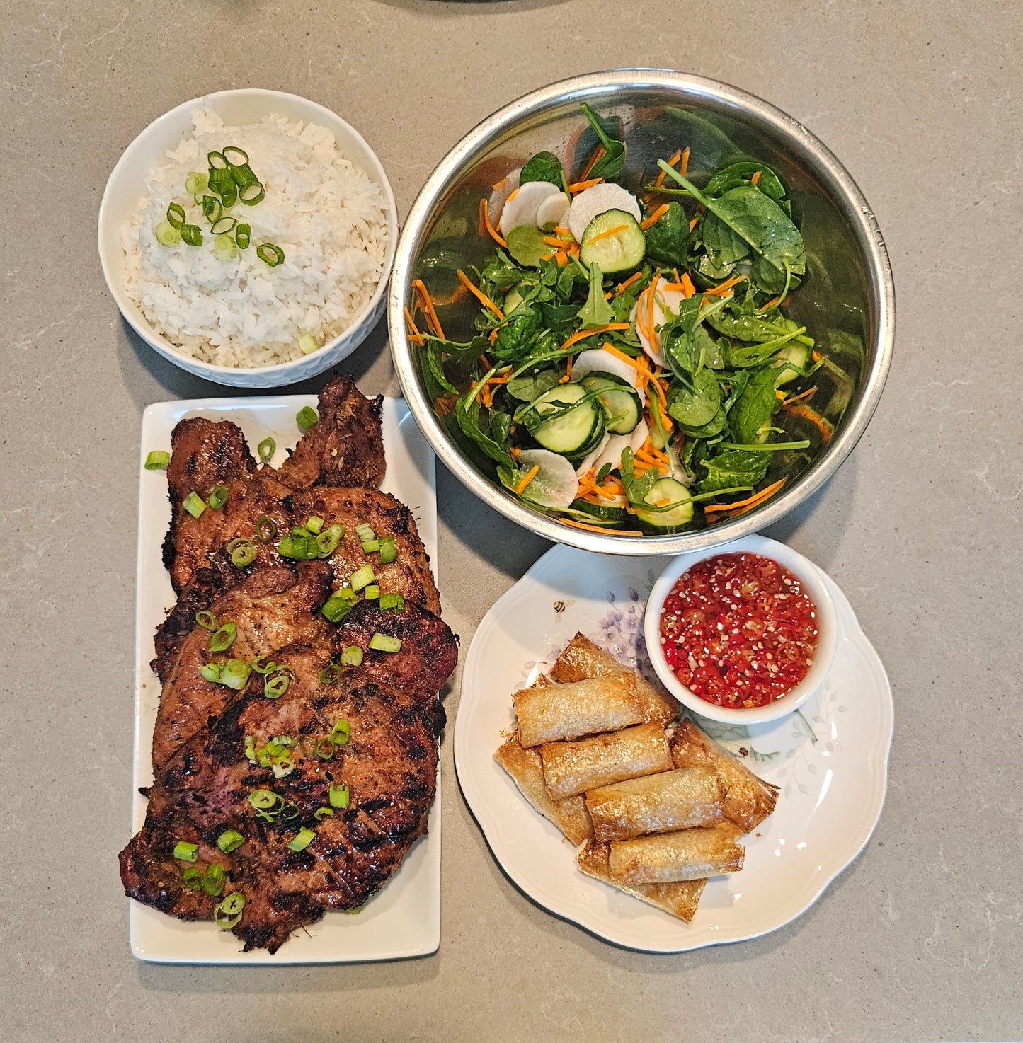 Grilled thai style pork chops on plate with bowl of salad white rice bowl and egg rolls with dipping sauce all on a gray table