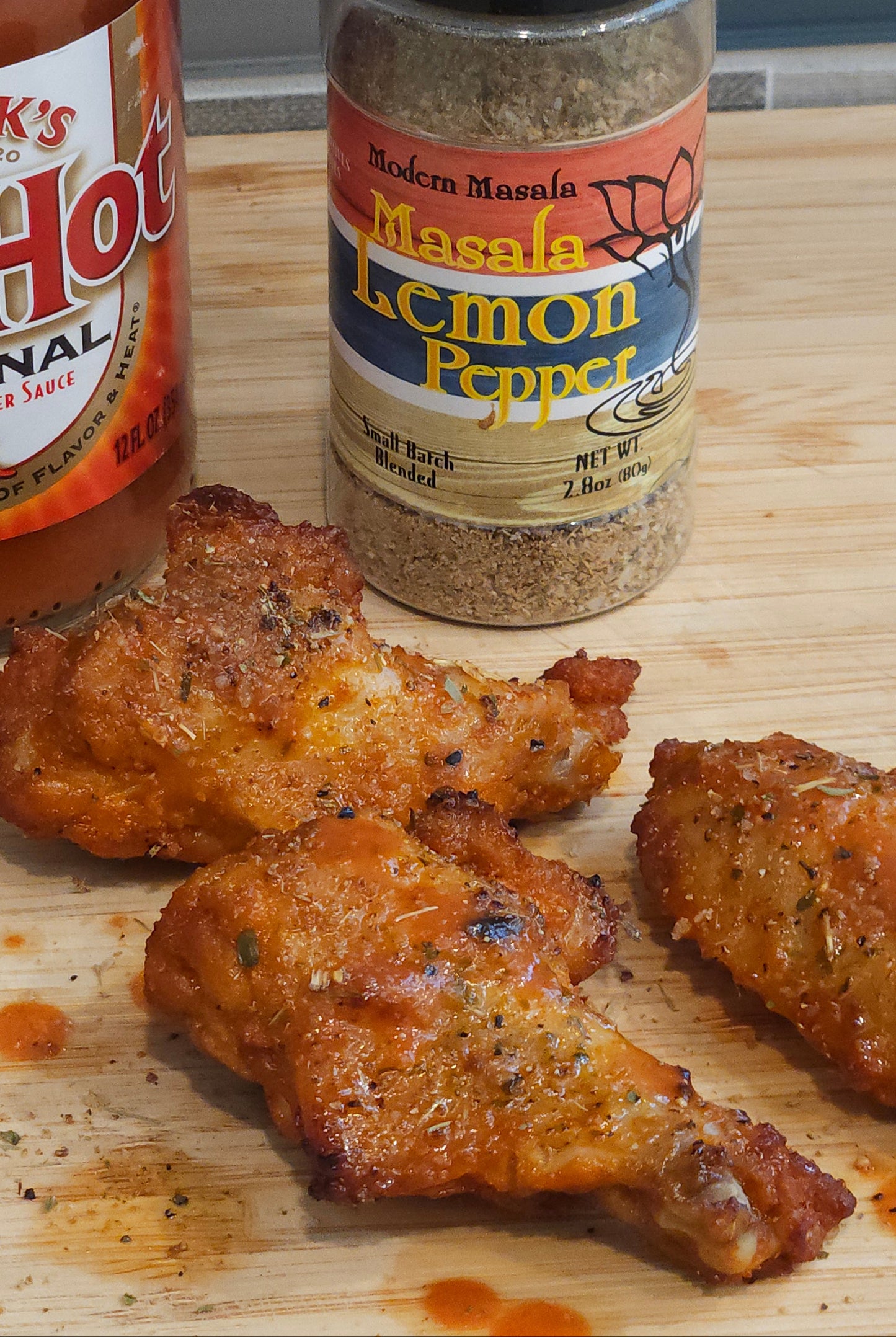 Spice bottle labeled 'Lemon Pepper' next to chicken wings on a wooden surface