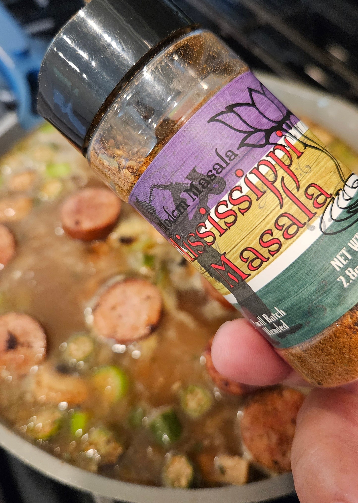 Hand holding a bottle of seasoning over a pot of cajun gumbo on a stove