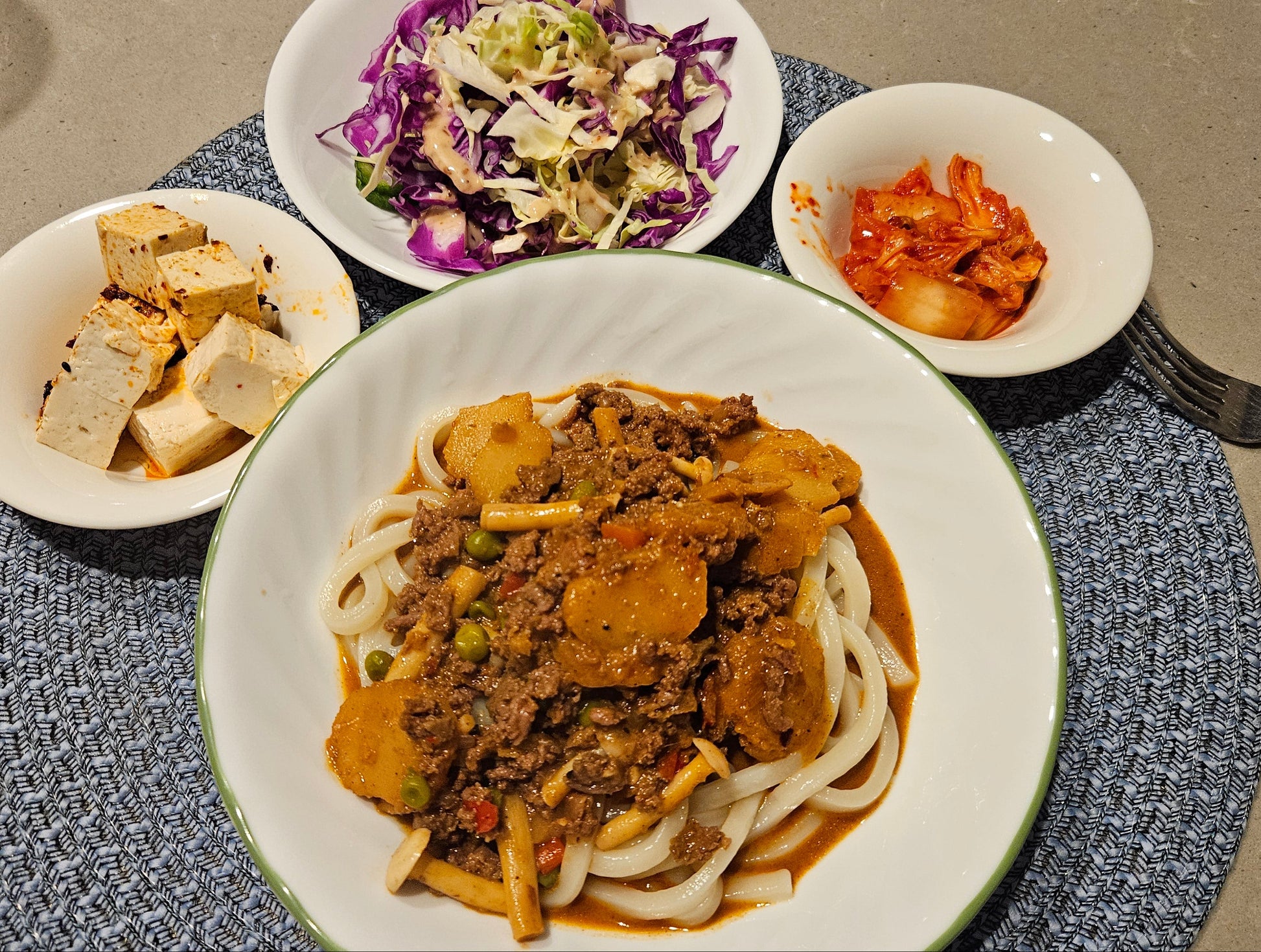 Plated dish of noodles with meat and vegetables, accompanied by side dishes on a textured placemat.